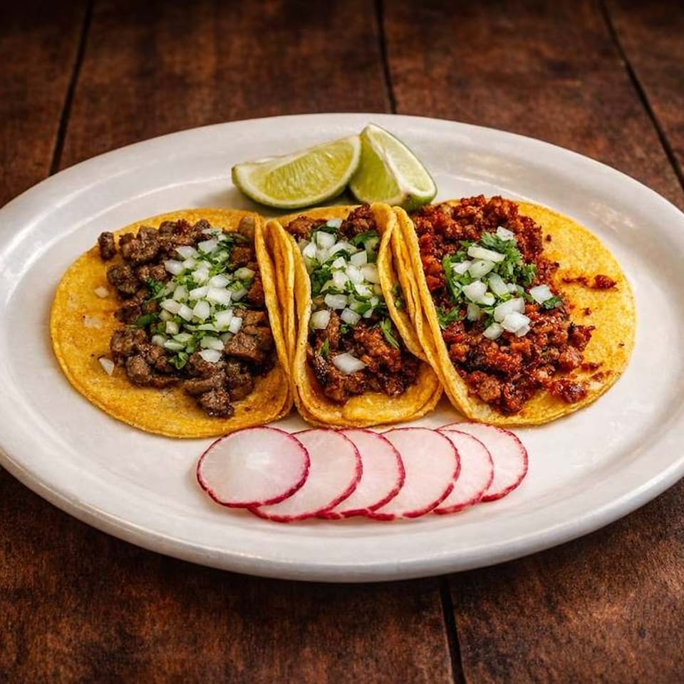 Three Mexican tacos topped with chopped onion and cilantro, served with lime wedges and sliced radishes on a white plate.