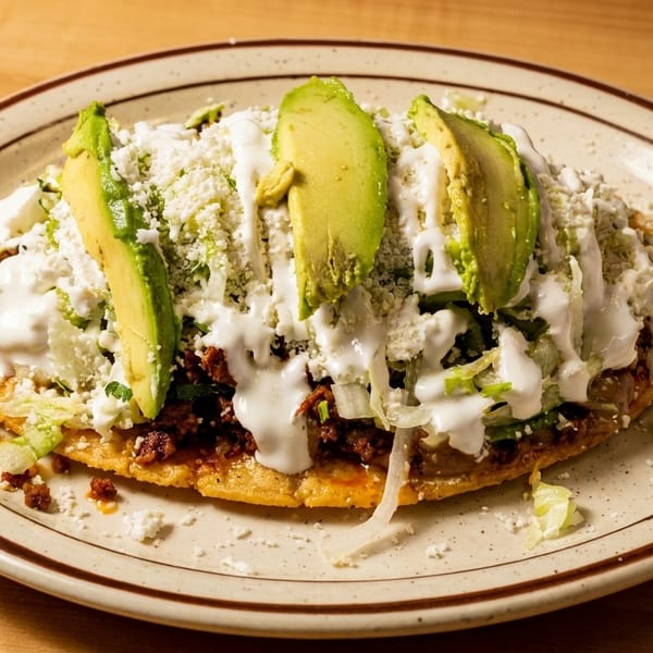 A Mexican sopes dish topped with meat, lettuce, cheese, sour cream, and avocado slices.