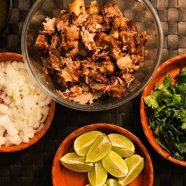 Overhead view of carnitas, chopped onions, cilantro, lime wedges, and salsa verde in bowls.