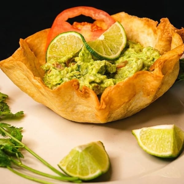 Guacamole in a crispy tortilla bowl, garnished with lime slices, tomato, and cilantro.