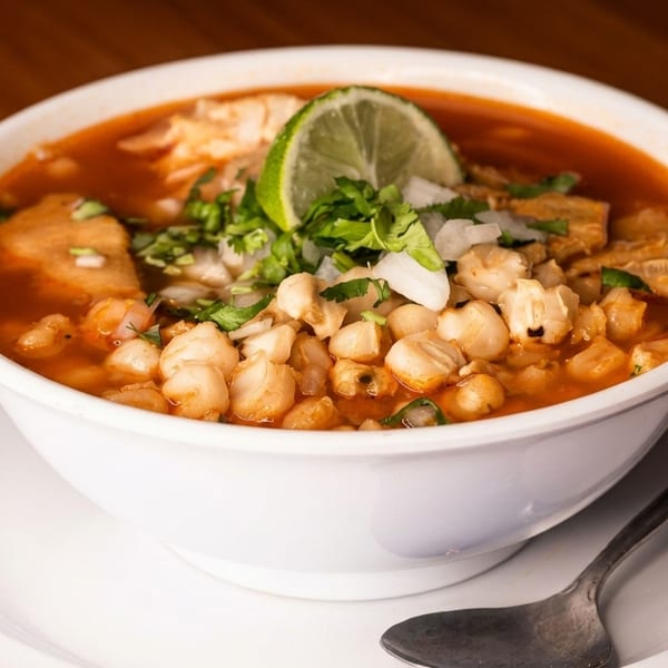 A bowl of menudo soup with hominy, tripe, cilantro, onions, and a lime wedge.