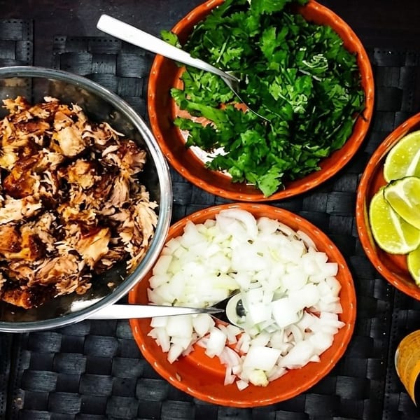 Overhead view of taco ingredients: shredded meat, cilantro, diced onions, lime wedges, and green salsa.