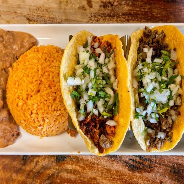 A white rectangular plate holds three tacos, refried beans with cheese, and orange rice.
